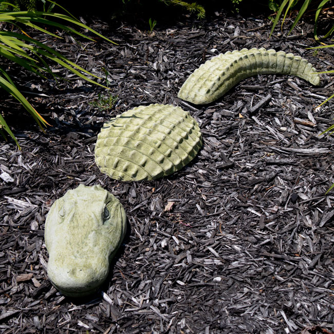 A three-piece moss green gator statue arranged in a garden bed with mulch, creating the illusion of an alligator emerging from the ground.